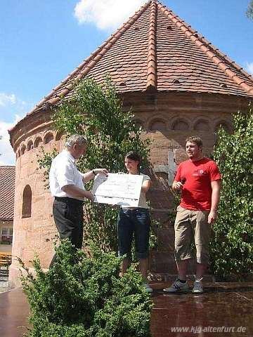 Die Pfarrjugendleiter Lisa und Christian übergeben Pfarrer Schiele beim Pfarrfest einen großen Scheck, im Hintergrund die Altenfurter Rundkapelle
