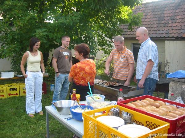 Andreas und Markus braten Würstchen auf dem Grill, daneben steht ein Tisch mit Salaten und Brötchen