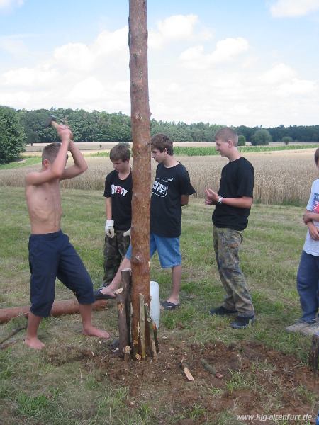 Ein Teilnehmer schlägt Holzstücke in den Boden, um die Standfestigkeit des Fahnenmasts zu verbessern