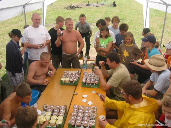 Stefan, Jürgen, Martin und Christian sitzen um zwei Biertische, jeder hat eine Palette Sahnepudding vor sich, rundherum stehen Teilnehmer und andere Leiter, die sie anfeuern, filmen und fotografieren