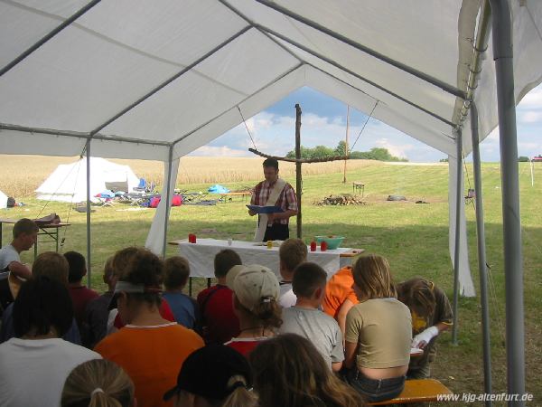 Gottesdienst unter dem Pavillon: Pastoralpraktikanten Peter Mairhofer steht hinter einem Altar aus einem Biertisch, dahinter hängt ein Holzkreuz