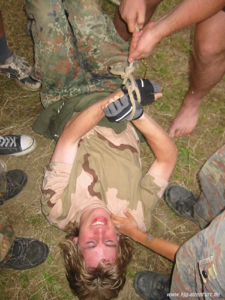 Ein Teilnehmer mit Tarnkleidung und Handschuhen liegt auf dem Boden und wird mit einem Seil gefesselt (natürlich nur als Spiel)