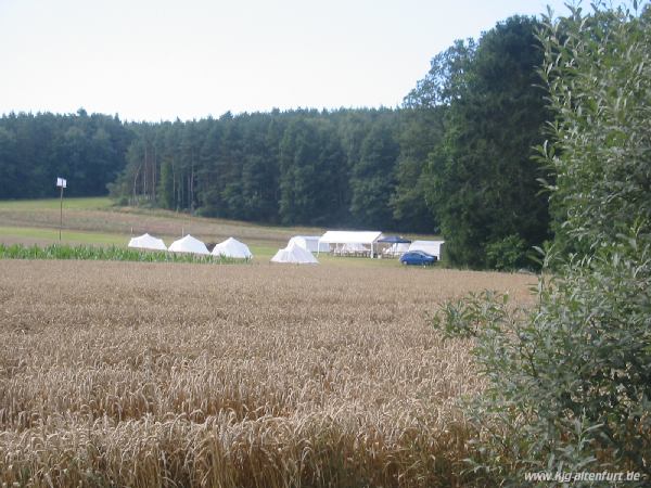 Im Fordergund ist ein Getreidefeld zu sehen, dahinter liegt unser Zeltplatz mit neun Zelten, einem kleinen und einem großen Pavillon, Fahne ...